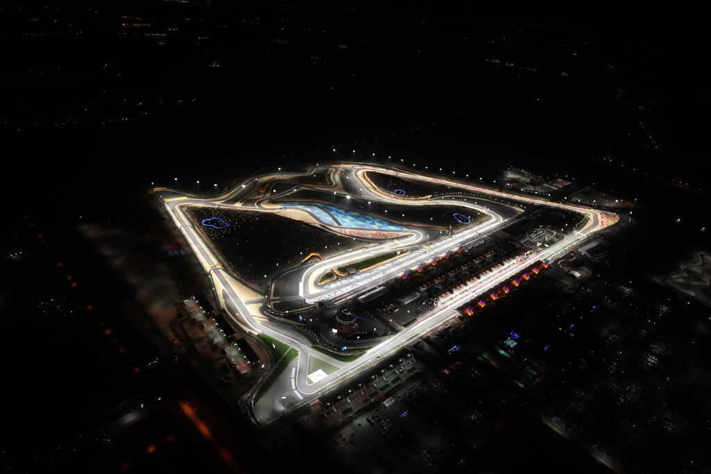 Overhead aerial view of the Bahrain Grand Prix at night showing Bahrain International Circuit illuminated under floodlights in the desert.