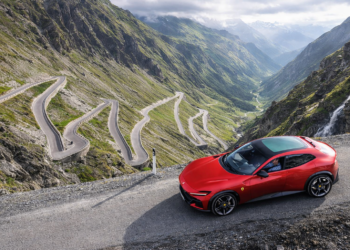 Red Ferrari-style luxury crossover parked on a dramatic alpine mountain road above a series of tight winding switchbacks and steep green valleys. Title: