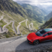 Red Ferrari-style luxury crossover parked on a dramatic alpine mountain road above a series of tight winding switchbacks and steep green valleys. Title: