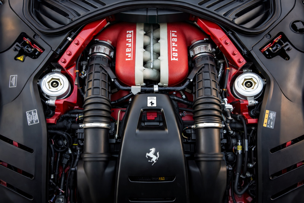 Close-up view of a high-performance red V12-style engine bay with black intake tubes, metallic hardware, and sculpted engine covers.