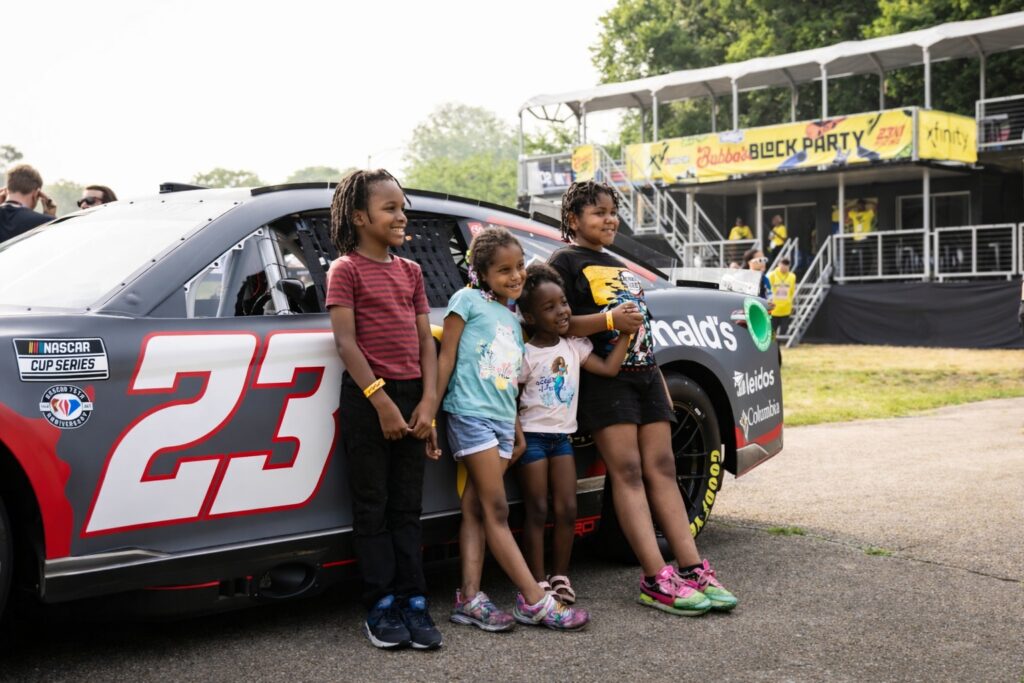 children posing beside Bubba Wallace number 23 NASCAR Cup Series car at community block party event