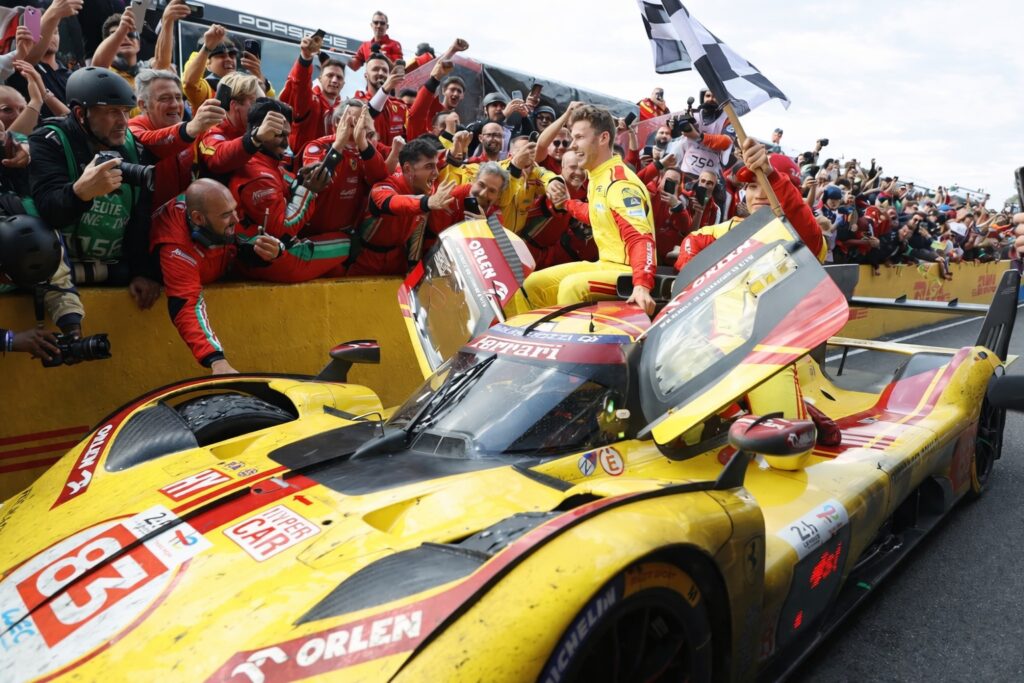 Winning endurance race driver celebrates from a yellow prototype race car while the team cheers and waves a checkered flag in pit lane.