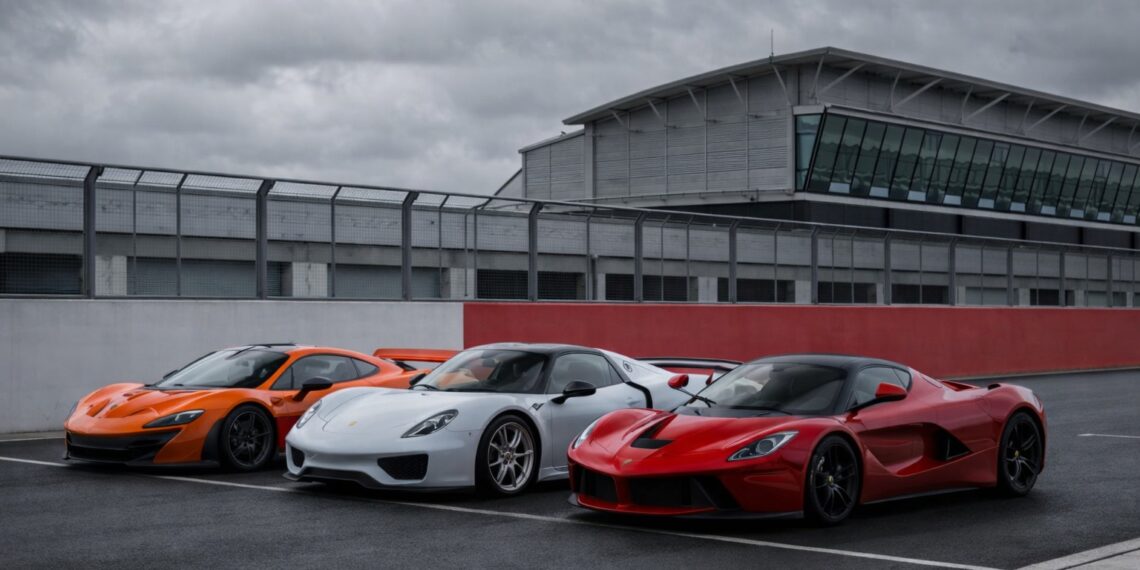 McLaren P1, Porsche 918 Spyder, and Ferrari LaFerrari lined up on a racetrack under an overcast sky