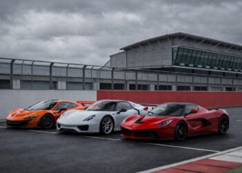 McLaren P1, Porsche 918 Spyder, and Ferrari LaFerrari lined up on a racetrack under an overcast sky