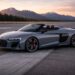 Metallic gray Audi R8 Spyder parked on an open runway at sunset with mountains and a pine tree backdrop
