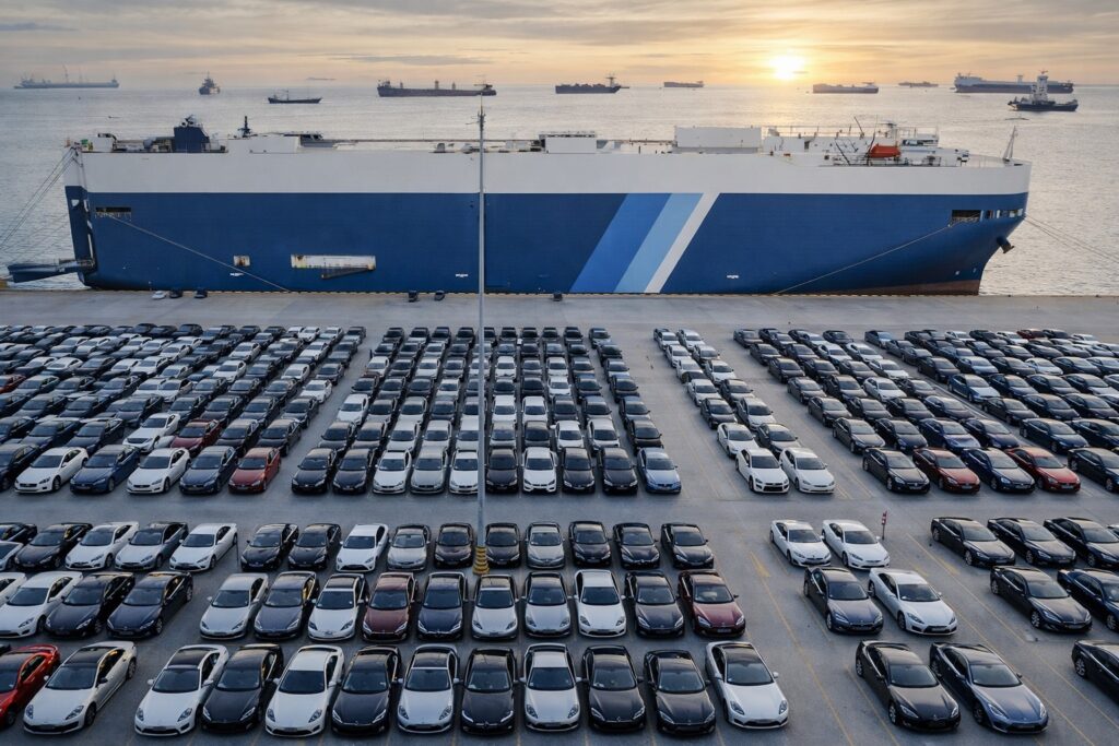 Luxury cars parked at a shipping port beside a RoRo cargo ship with multiple vessels visible on the water at sunset