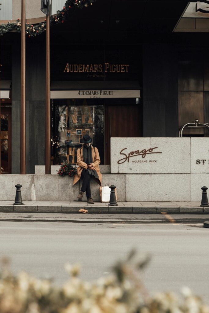 Man in a camel coat and hat sitting outside a luxury watch boutique storefront on a city street
