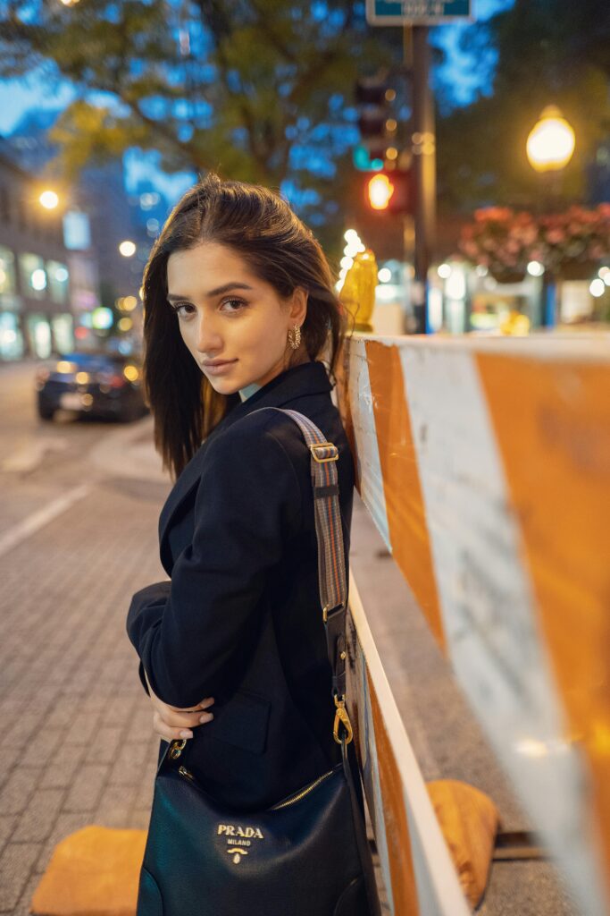 Woman in black blazer standing on a city street at dusk carrying a luxury shoulder bag with gold accents