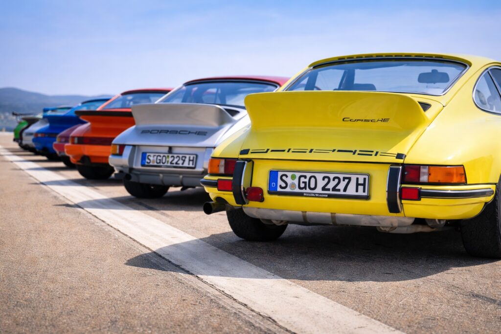 Classic Porsche 911 Carrera RS lineup in multiple colors parked on airstrip with coastal backdrop