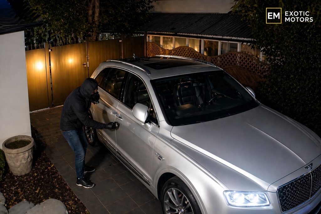 Masked person attempting to break into a silver luxury SUV parked in a driveway at night
