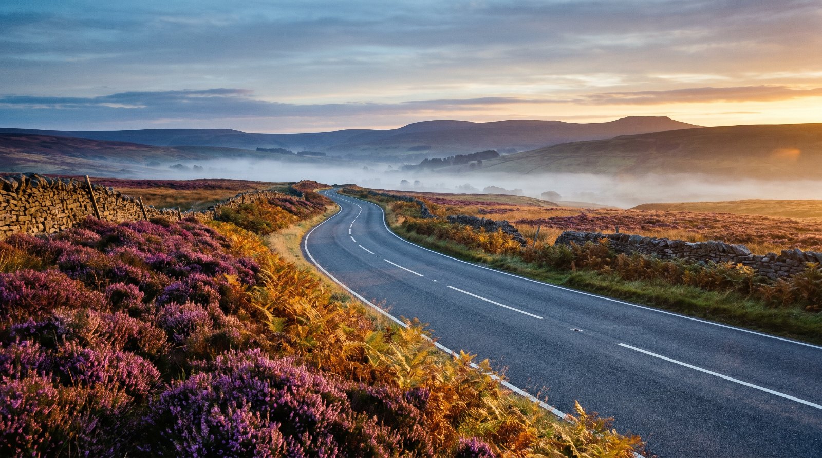 Empty two-lane road sweeping through British moorland at dawn with dry stone walls and low valley mist