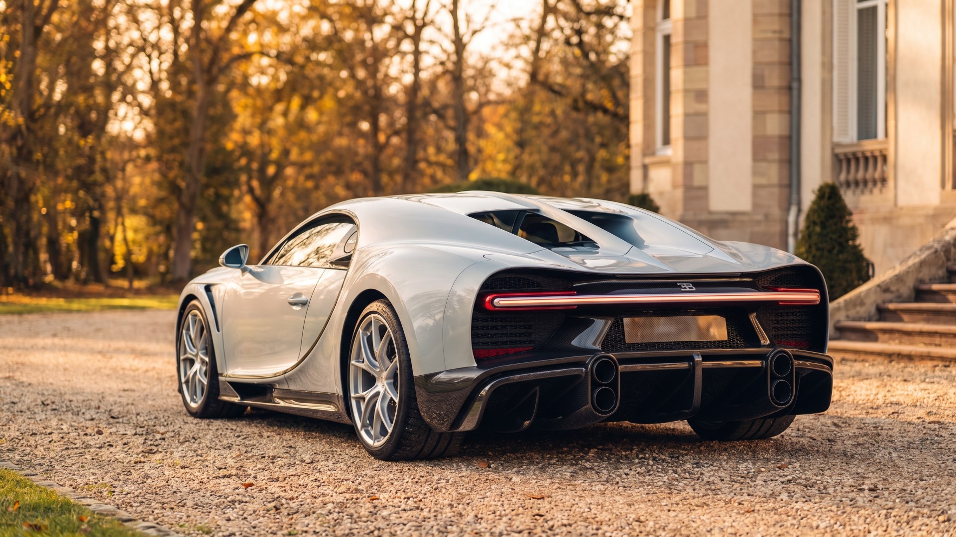Silver Bugatti Chiron seen from the rear three-quarter on a gravel château drive in autumn light, the W16-era car of Bugatti's Volkswagen years. AI-generated editorial visualization, not a photograph.