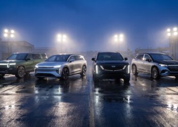 Four electric super-SUVs lined up with headlights on across a wet airfield apron at dusk: green Rivian R1S, silver Lucid Gravity, black Cadillac ESCALADE IQ and two-tone Mercedes-Maybach EQS SUV