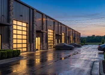 Row of modern garage condo units with tall glass overhead doors lit from within at dusk