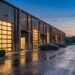 Row of modern garage condo units with tall glass overhead doors lit from within at dusk