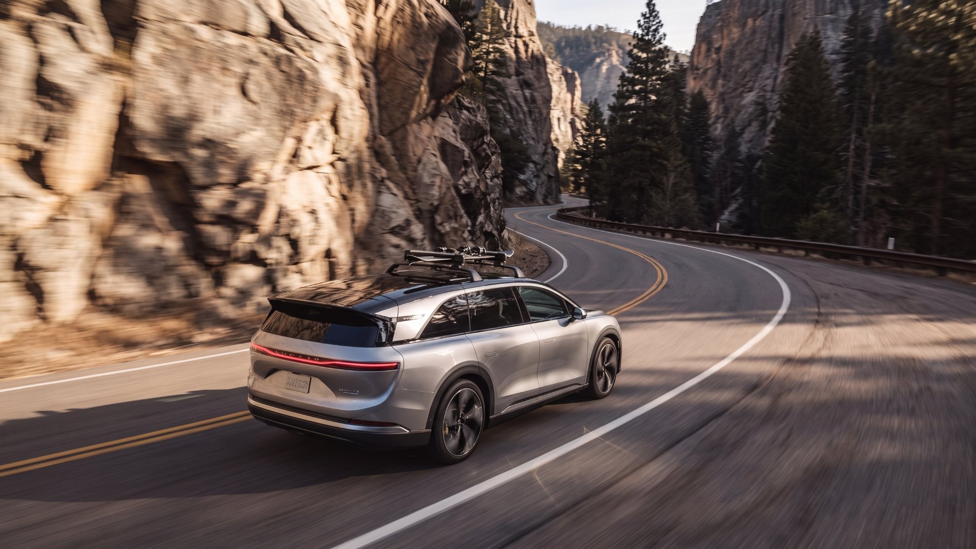 Silver Lucid Gravity driving through a curve on a mountain canyon road between rock walls and pine trees