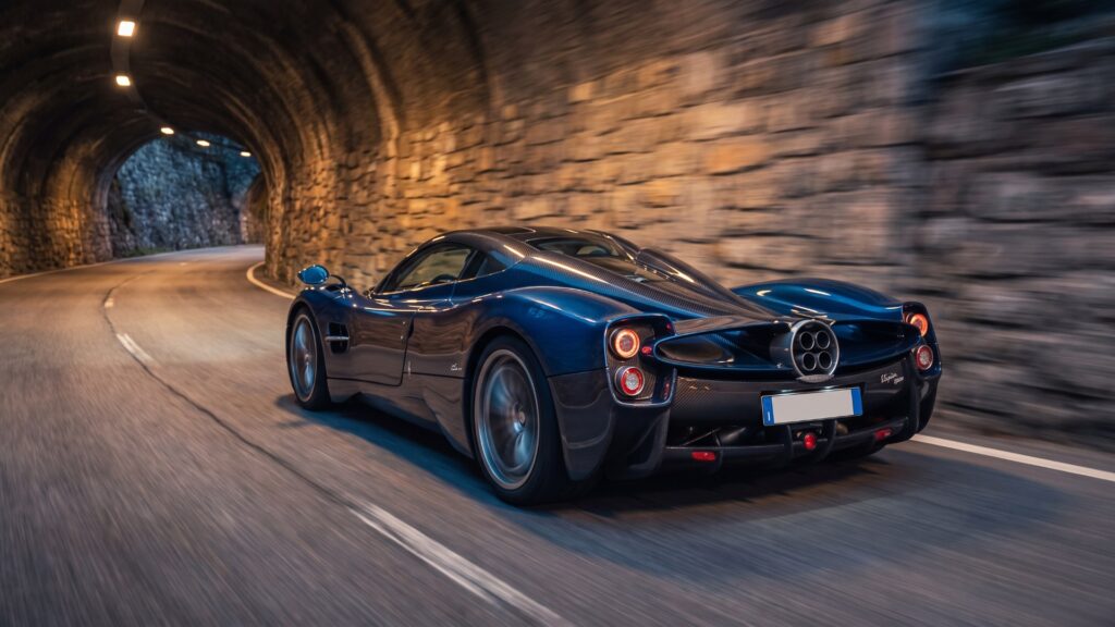 Blue Pagani Utopia hypercar driving through a lit stone mountain tunnel at dusk, its quad exhaust visible
