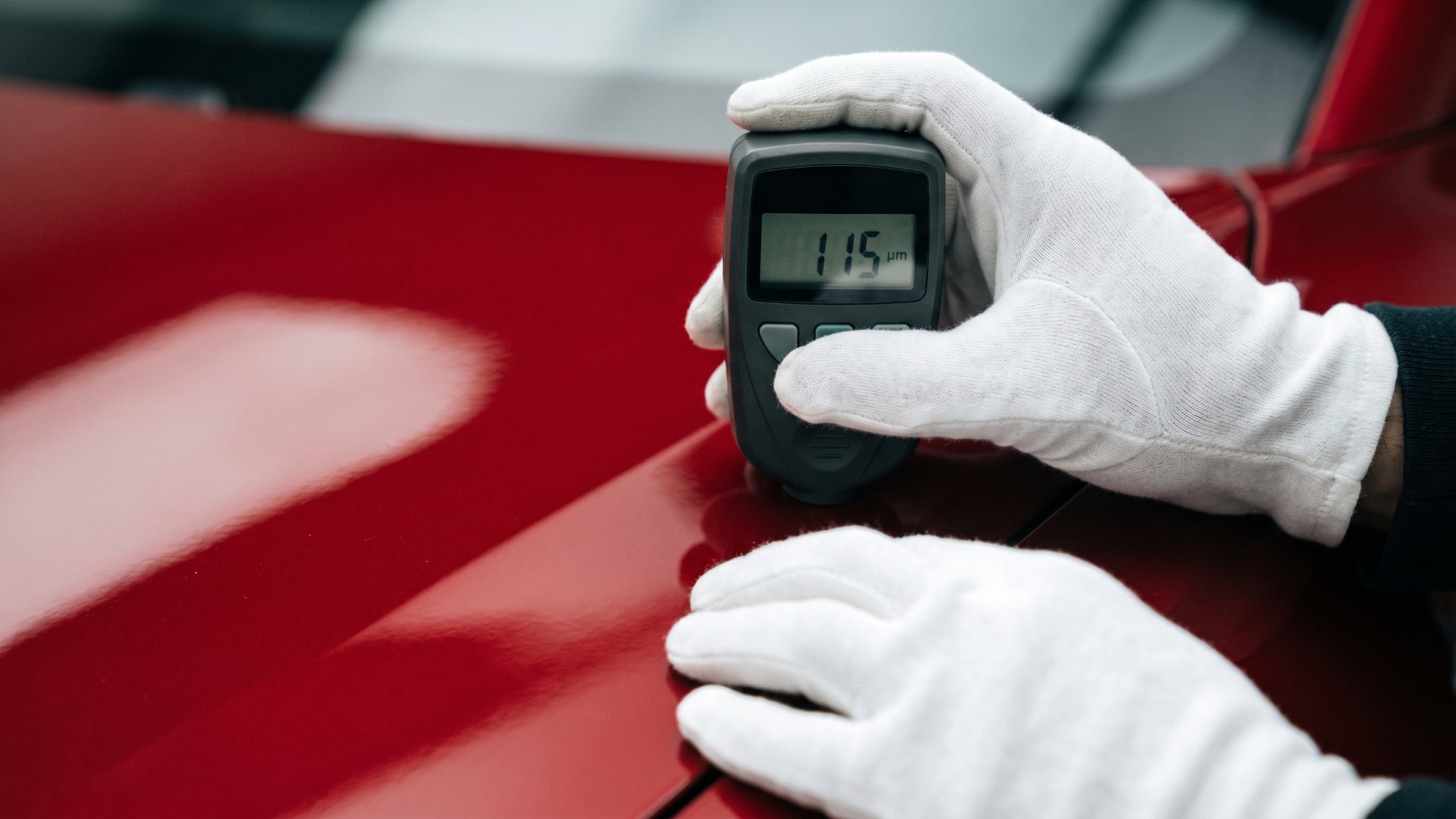 A gloved hand holding a digital paint-thickness gauge against a deep red body panel during a condition inspection