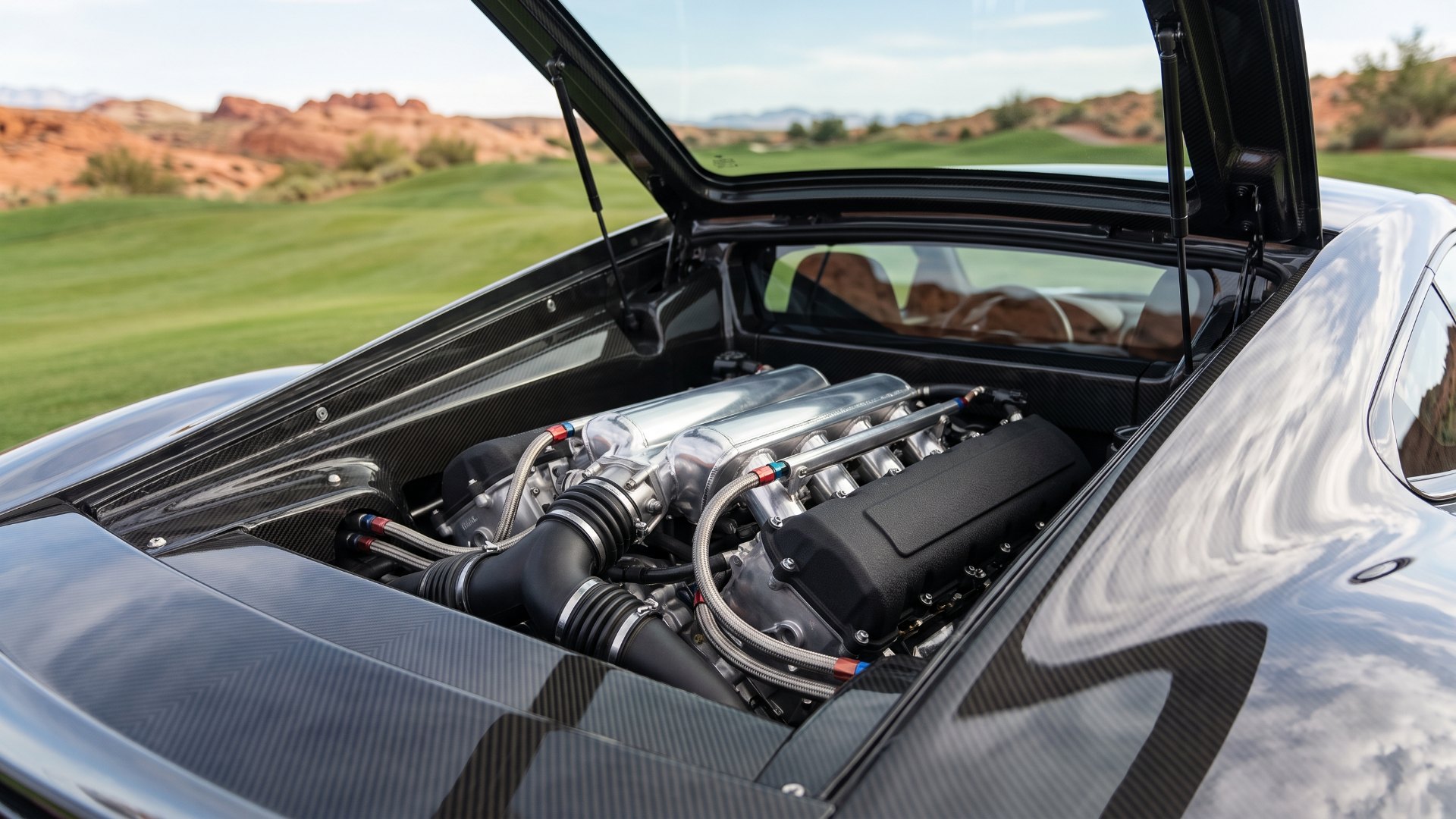 Close-up of the open glass engine cover of a modern mid-engine supercar showing a naturally aspirated V12 with polished intake plenums and carbon fibre surrounds