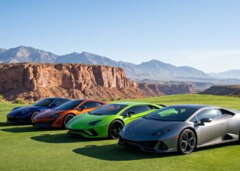 A row of modern supercars parked on manicured golf course fairway grass with red sandstone cliffs and the Spring Mountains behind them
