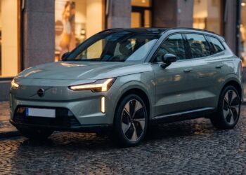 Sage-grey Volvo EX60 electric SUV parked on a wet cobblestone street at dusk