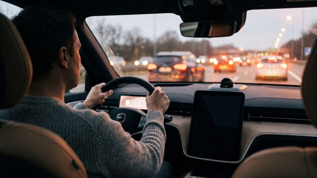 Driver with both hands on the wheel of a Volvo EX90 on a highway at dusk, with a cabin camera on the dashboard