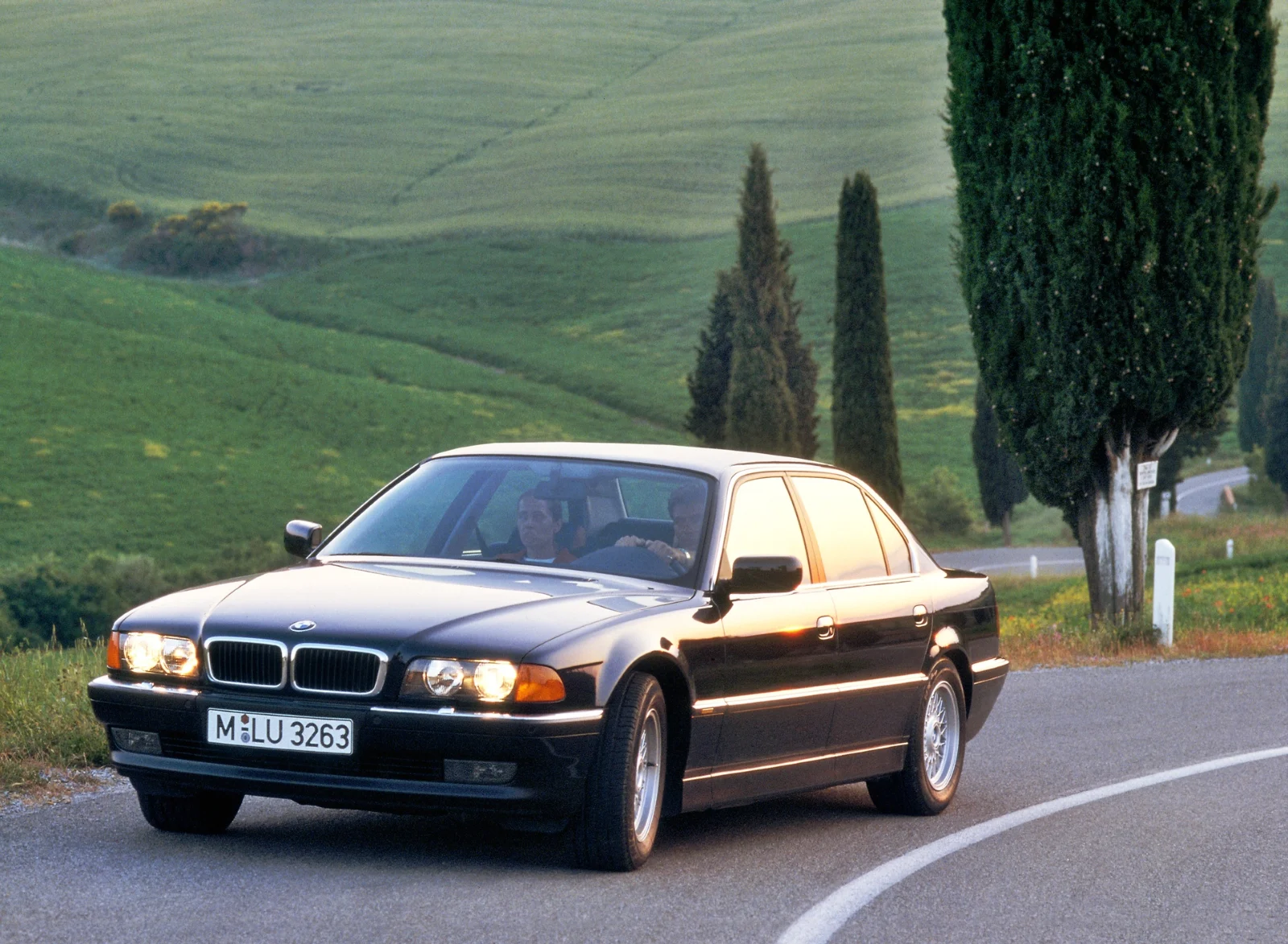 Black BMW 7 Series E38 sedan driving on a country road with cypress trees