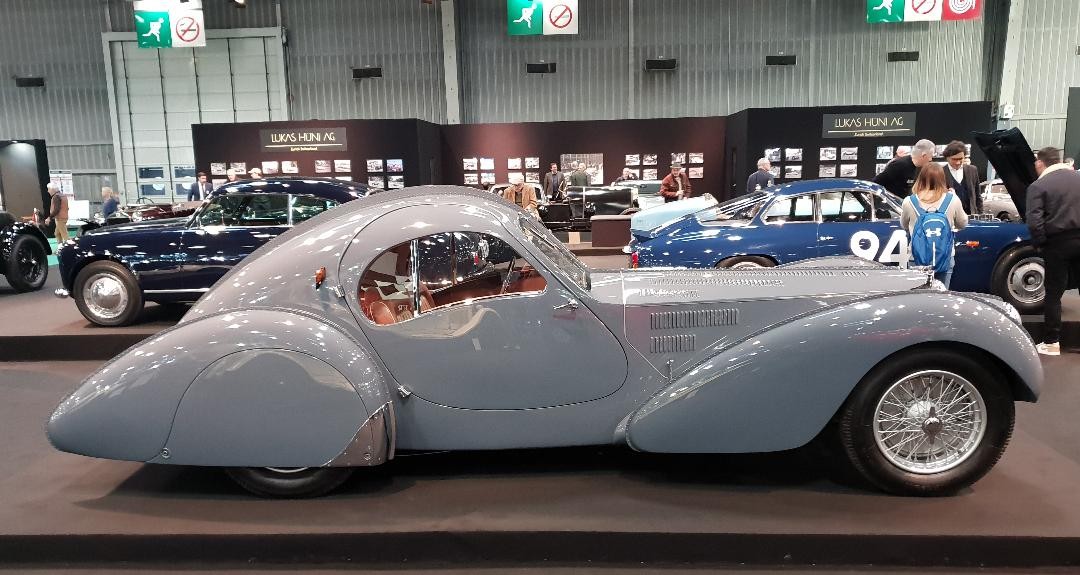Side view of a blue-grey 1936 Bugatti Type 57 SC Atlantic coupe, chassis 57473, with its riveted dorsal seam, at an indoor classic car show