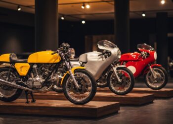 Yellow, silver and red 1970s sport motorcycles displayed on wooden plinths in a dim auction preview hall