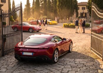 A dark red Ferrari Roma waits at open estate gates while owners gather around classic and modern Ferraris in the courtyard beyond