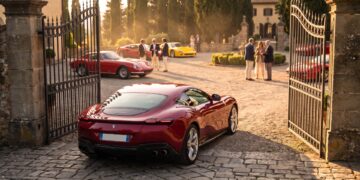 A dark red Ferrari Roma waits at open estate gates while owners gather around classic and modern Ferraris in the courtyard beyond
