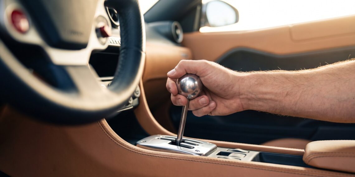 Driver’s hand on a polished aluminum gear knob above an open metal shift gate in a tan leather cabin