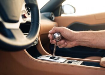 Driver’s hand on a polished aluminum gear knob above an open metal shift gate in a tan leather cabin