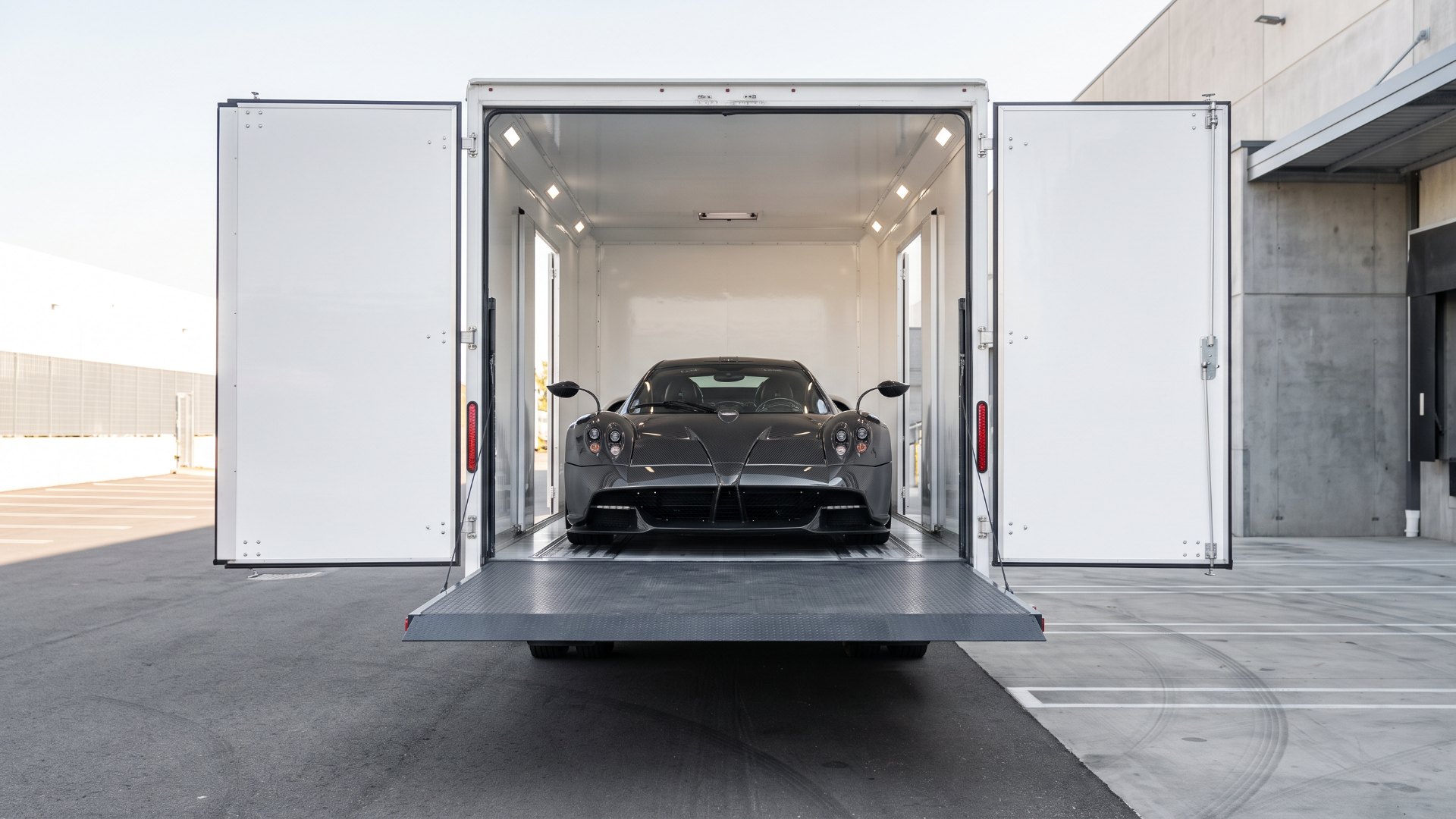 Illustration of a carbon-fiber hypercar resembling a Pagani Huayra inside a hard-sided enclosed transport trailer, with the rear hydraulic liftgate raised level with the trailer floor