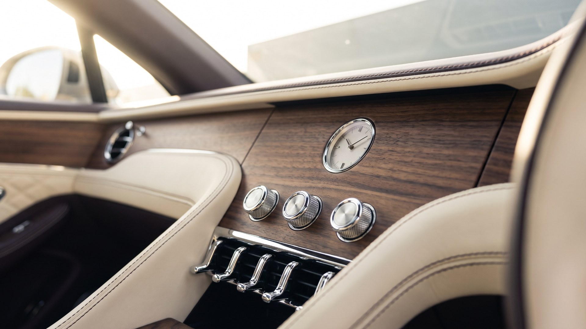 Small analog clock set into a walnut dashboard above knurled metal knobs and chrome vent pulls in a cream leather luxury cabin