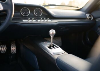 Illustration of a polished metal gear lever in an open gate and three drilled pedals in a minimalist hypercar cabin with two analog gauges
