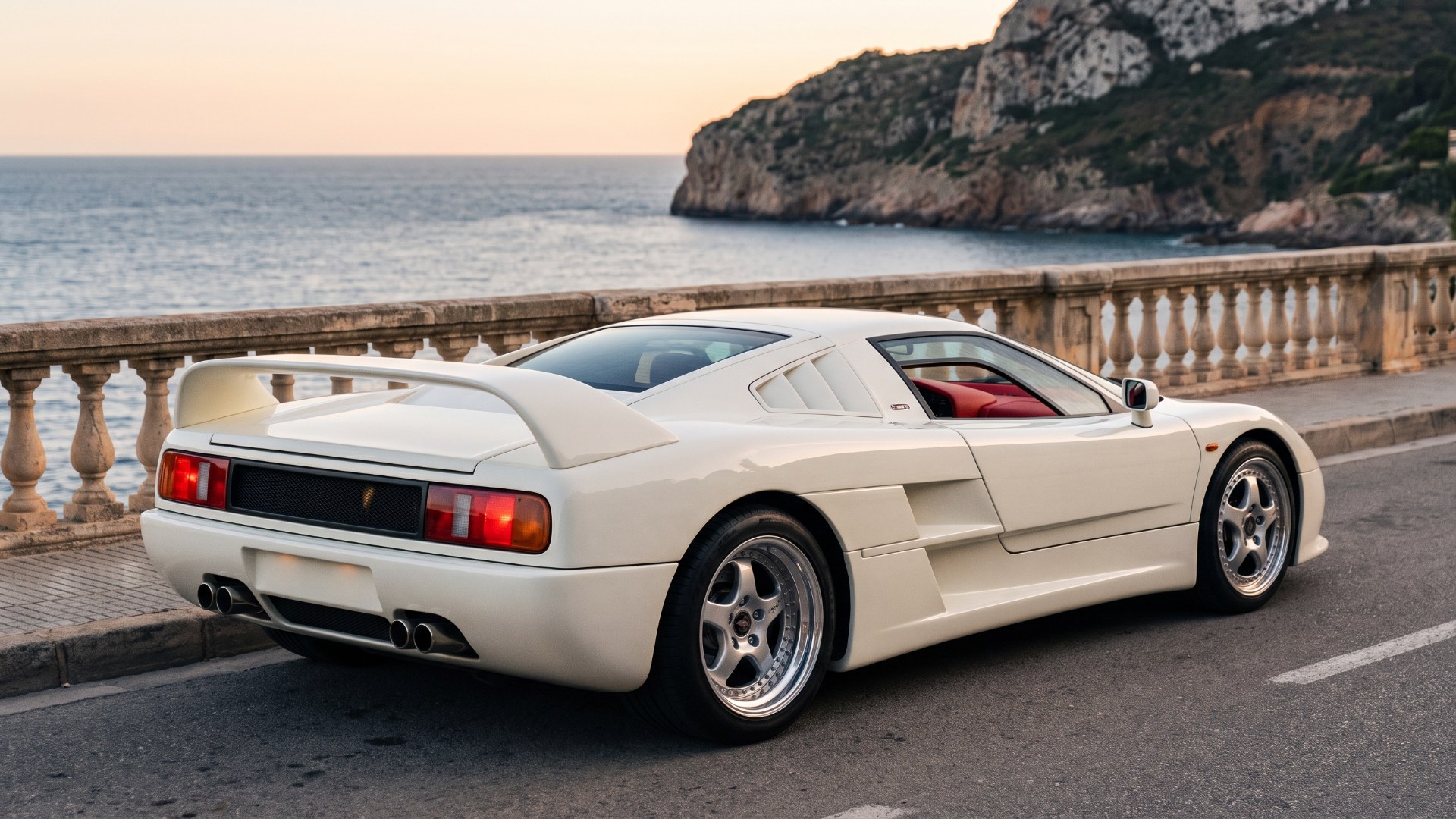 Rear three-quarter view of an ivory-white MCA Centenaire on a coastal road at sunset