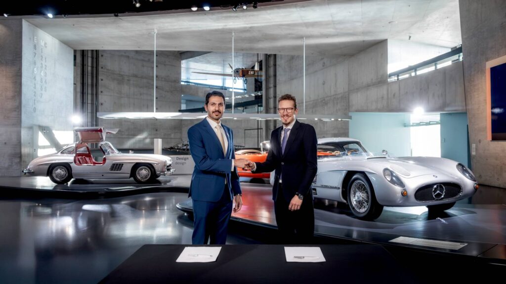 Executives from Binghatti and Mercedes-Benz shake hands at a signing table in the Mercedes-Benz Museum, with a silver 300 SL Gullwing with open doors behind them