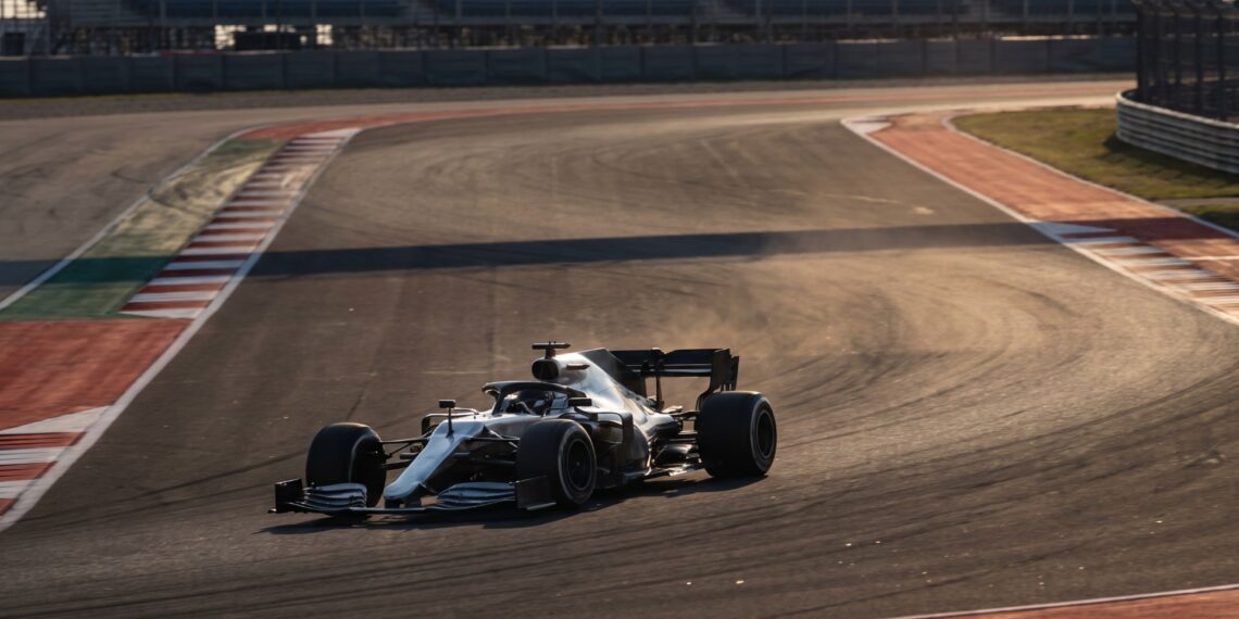 Illustration of an unbranded silver Formula One car cornering on a sunlit race circuit with empty grandstands behind