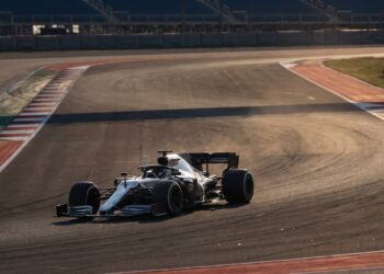 Illustration of an unbranded silver Formula One car cornering on a sunlit race circuit with empty grandstands behind