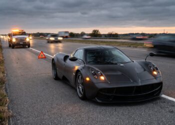 Illustration of a dark carbon-fiber hypercar resembling a Pagani Huayra stopped on an interstate shoulder at dusk with hazard lights on, a warning triangle behind it and a recovery truck approaching in the distance