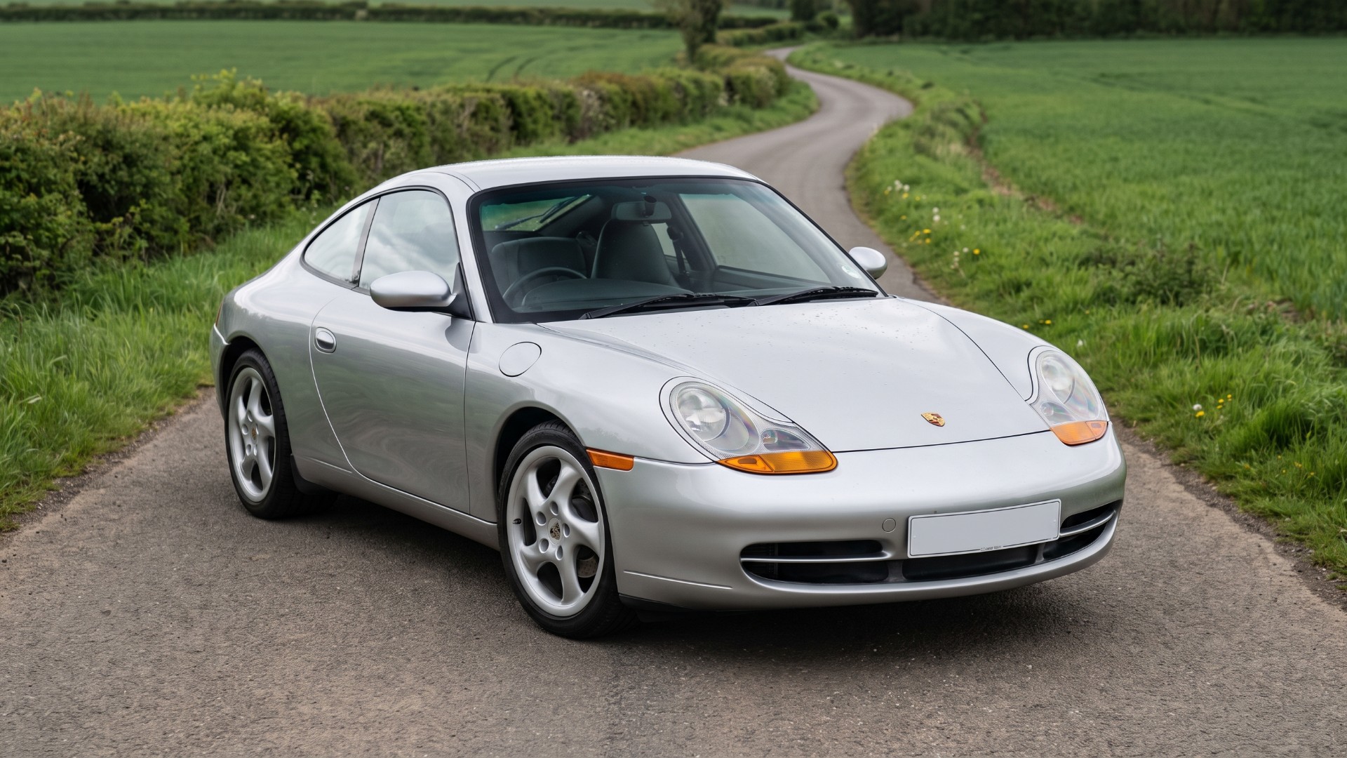 Silver 996-generation Porsche 911 Carrera on a country lane, showing its one-piece headlights with amber indicators