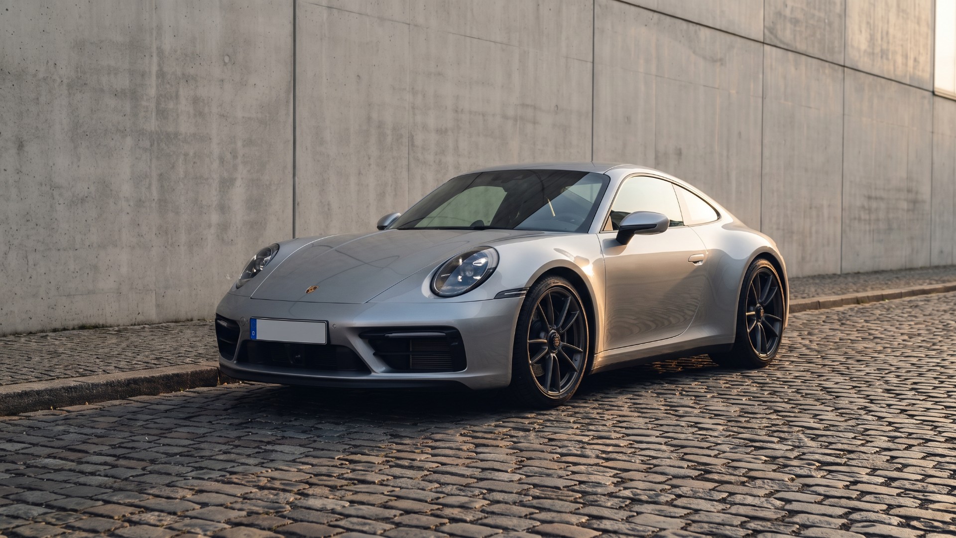 Silver Porsche 911 coupe parked on a cobbled street beside a concrete wall, seen from the front three-quarter