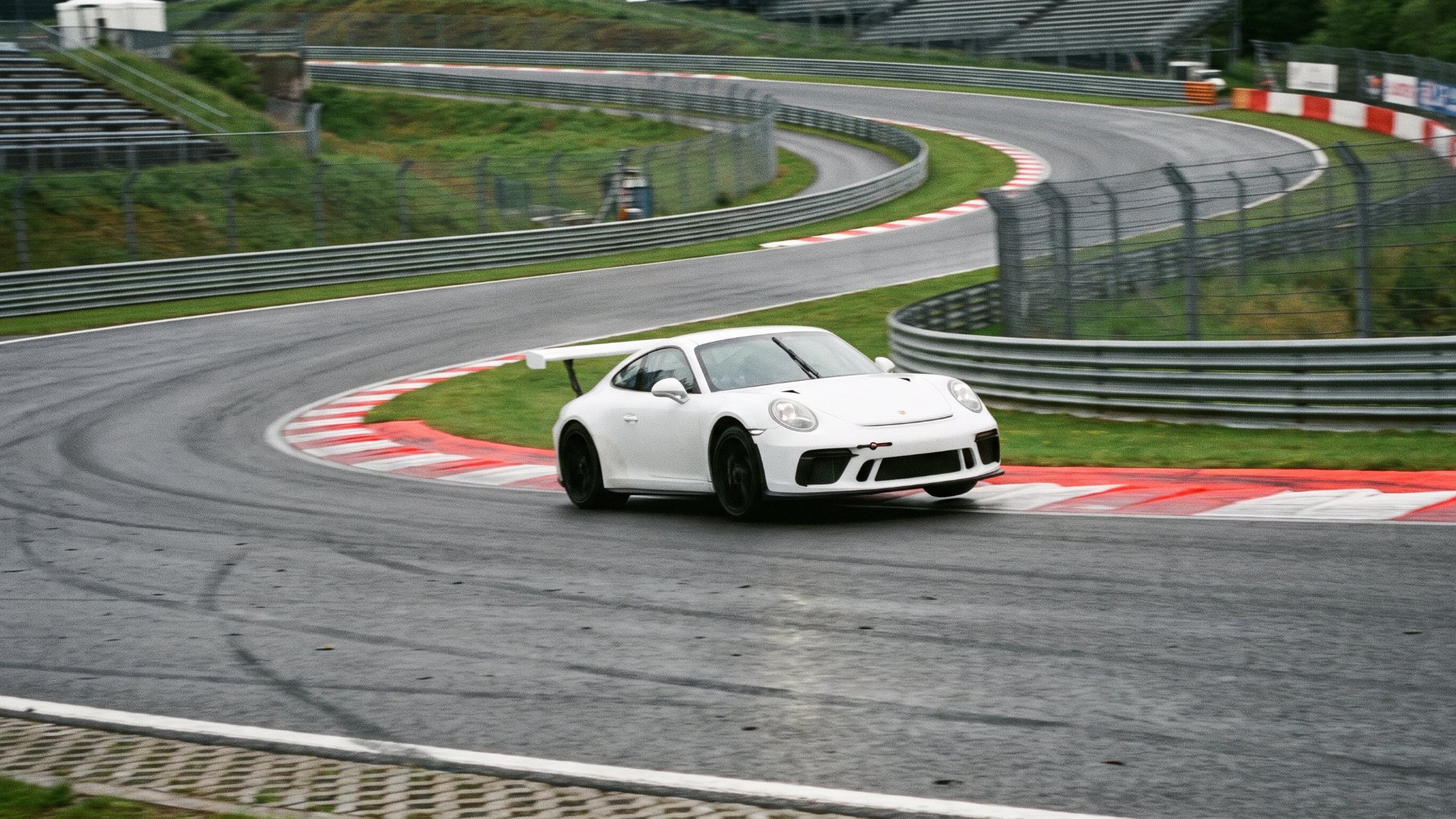 White Porsche 911 with a rear wing cornering over red-and-white curbs on a wet racing circuit
