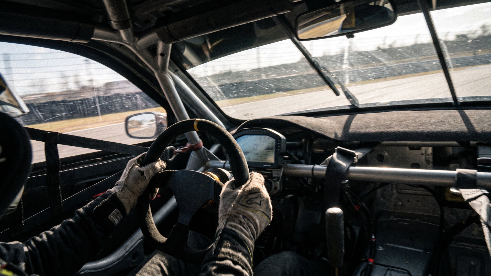Driver's gloved hands on a suede race car steering wheel inside a cockpit with a roll cage and digital dash