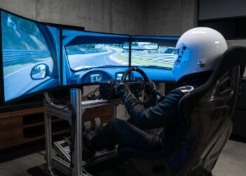 Sim racer in a white full-face helmet and race suit at a professional racing simulator with a direct-drive wheel and three curved screens showing a circuit