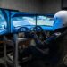 Sim racer in a white full-face helmet and race suit at a professional racing simulator with a direct-drive wheel and three curved screens showing a circuit