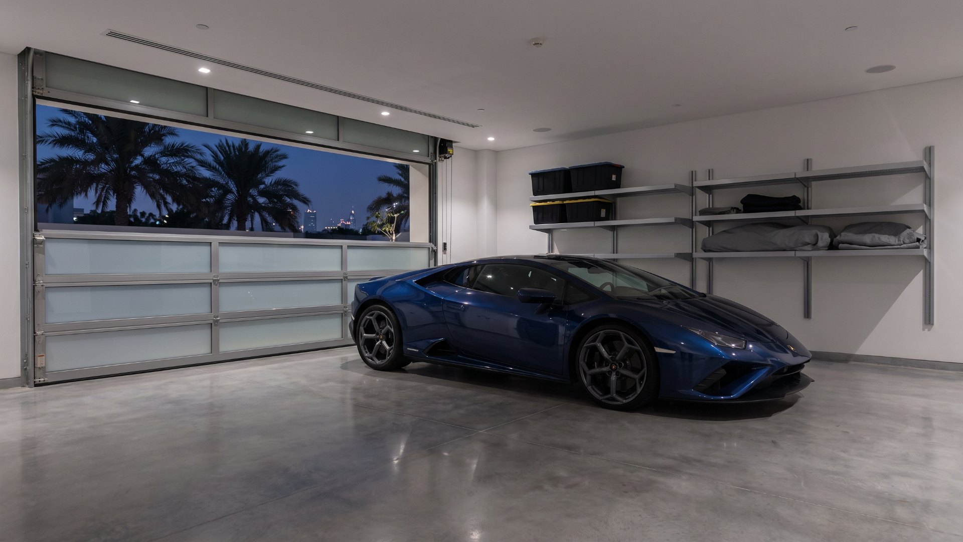 Dark blue mid-engine supercar parked in a modern private villa garage in Dubai at dusk, with palm trees visible through the partly open door