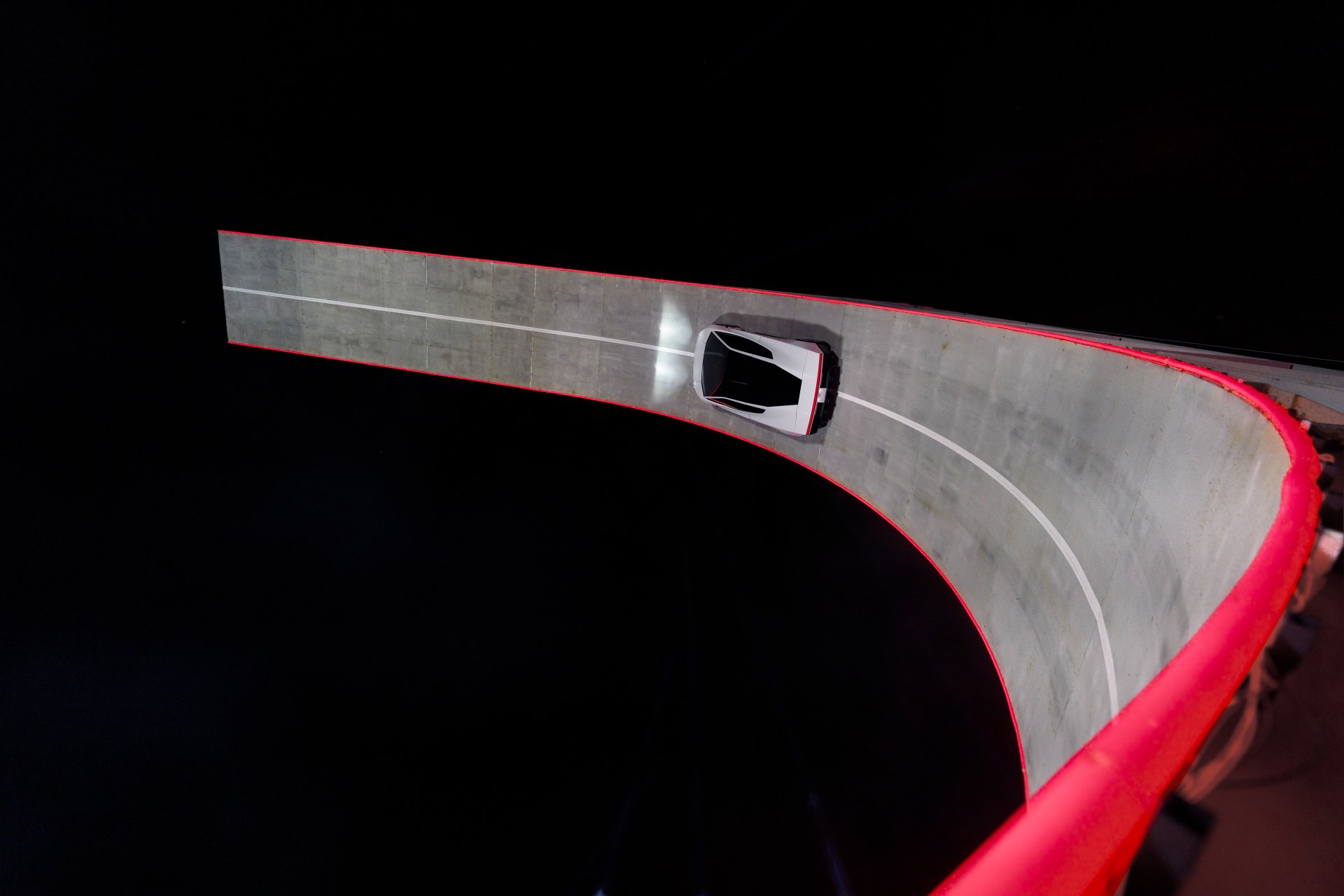 Tesla Roadster driving along a curved concrete ramp that turns inverted, seen from above at night