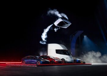 Tesla Roadster prototype airborne on thruster jets above a Tesla Semi and other Tesla vehicles at night